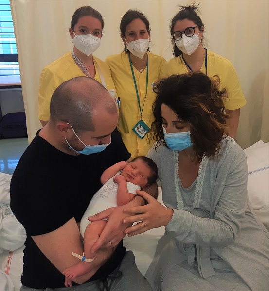 Nella foto, scattata in una stanza del reparto di ostetricia si vede il papà che tiene in braccio la neonata, la mamma con una mano sopra il braccio del papà in modo amorevole, sono seduti sul letto, dietro tre ostetriche in piedi 