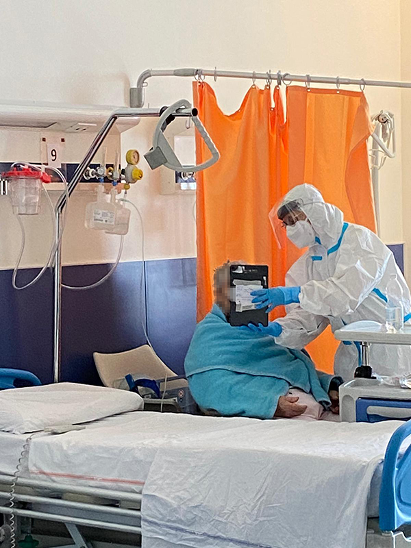 Nella foto si vede una persona ricoverata seduta su una poltrona e una operatore con le protezioni necessarie che sorregge un tablet per permettere una videochiamata