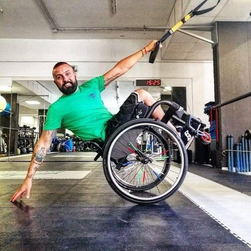 Nella foto Antonio all'interno di una palestra, sorridente, con la carrozzina alzata nella parte davanti con il braccio sinistro si regge a una corda e il braccio destro teso all'indietro con la mano che sfiora il pavimento. Allegria e voglia di vivere emergono dall'immagine 