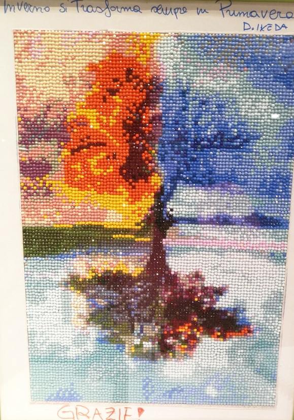 Nella foto il quadro realizzato dalla giovane donna con la tecnica del diamond painting. Nel quadro è raffigurato un albero con la chioma suddivisa a metà, la parte destra (rispetto allo spettatore) con colori caldi, arancione, giallo e un po' di marrone) la parte sinistra con colore azzurro, predominante, bianco e blu), le porzioni di cielo in secondo piano sono dalla parte destra: rosa, vinaccia  e avorio; dalla parte sinistra azzurro con una piccola parte di verde acqua.  Il fusto è di colore marrone. L'albero è riflesso in uno specchio d'acqua. La dedica riportata sul quadro è: nella parte alte, scritta di azzurro "L'inverno si trasforma sempre in primavera"  D. Ikeda. Nella parte in basso "Grazie!" scritto di rosso 